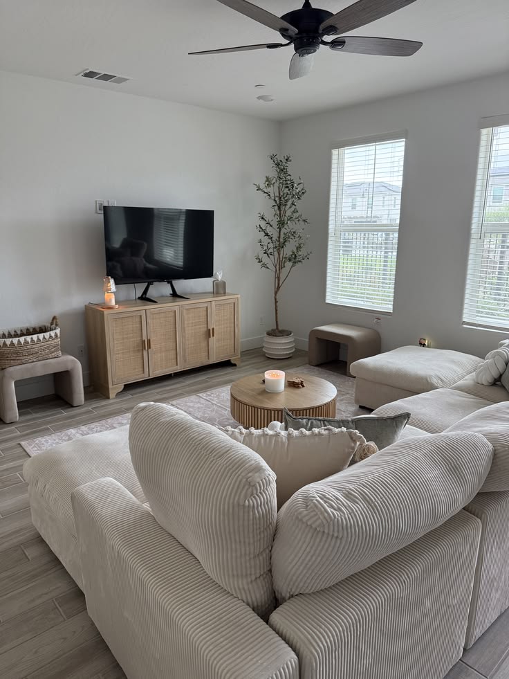 Bright living room with a cream corduroy sectional, rattan front media console with a TV, potted olive tree, round wooden coffee table, and large shuttered windows.