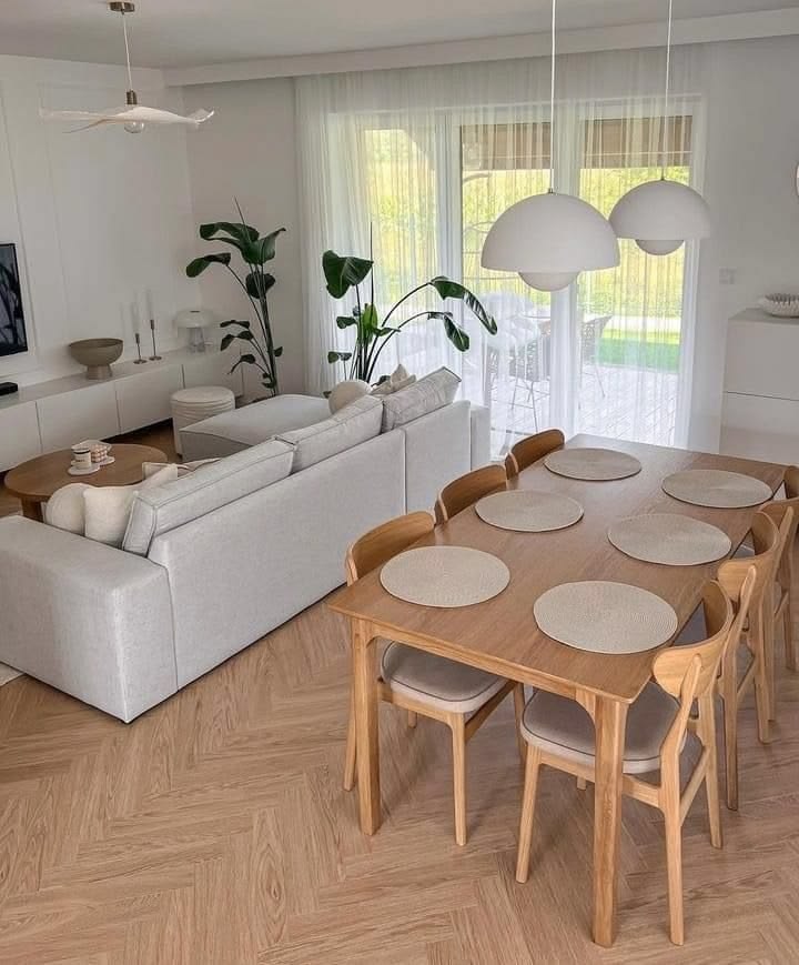 Open plan living and dining space with herringbone floors, a white sectional, low white media unit, oak dining table with pale chairs, dome pendant lights, and large plants.