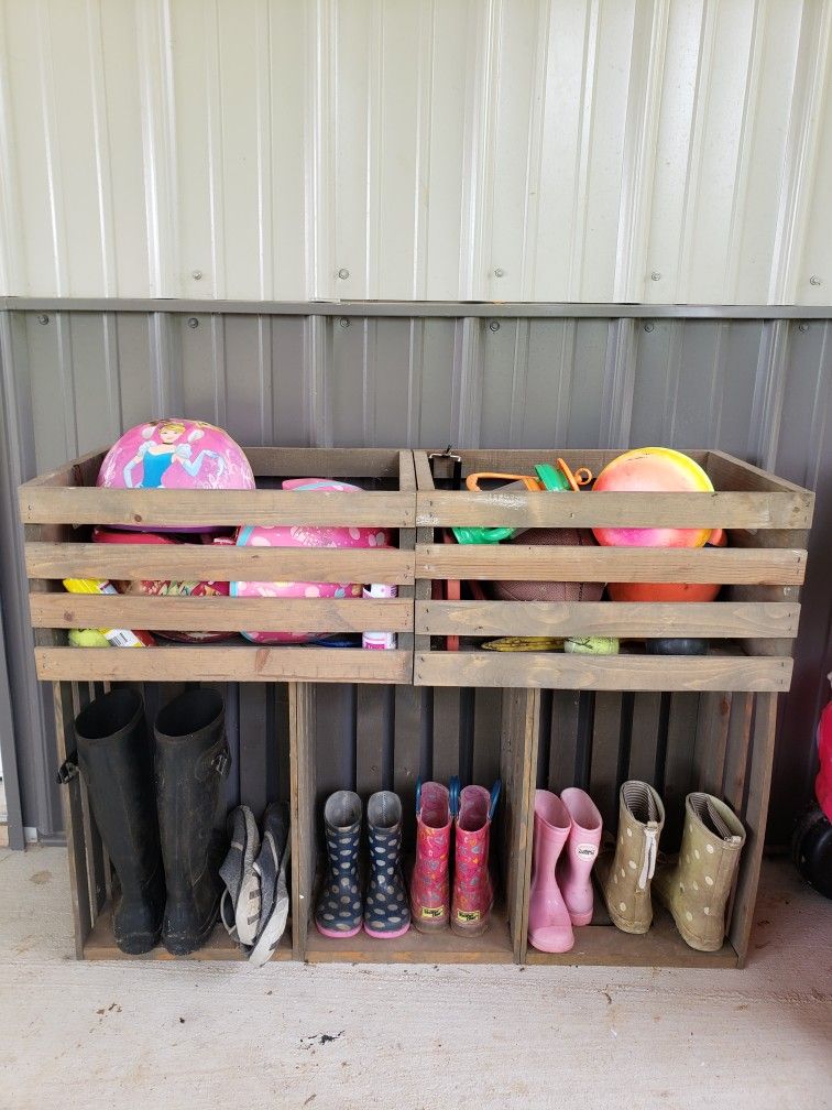 Wooden crate unit against a corrugated metal wall, with rain boots stored in the lower cubbies and balls and outdoor toys in the crates above.