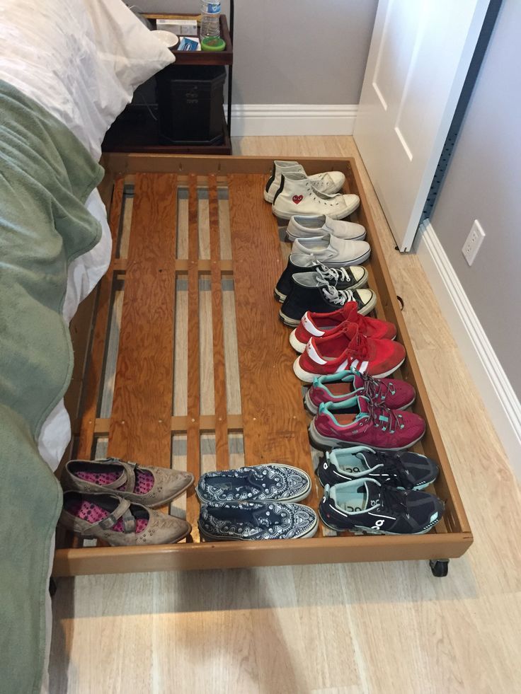 Wooden rolling shoe tray on casters pulled out from under a bed, holding sneakers and flats arranged in rows.