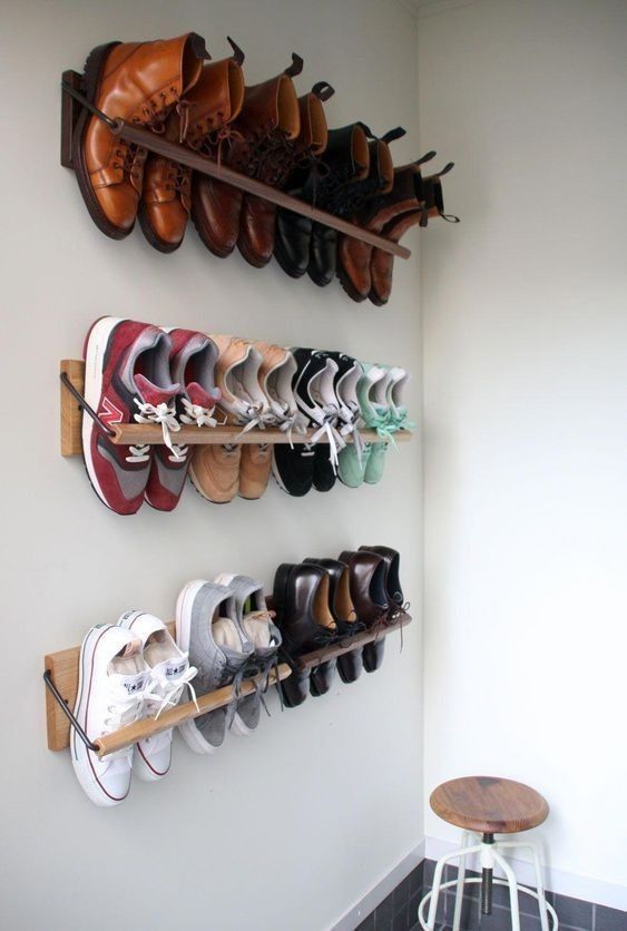 Three wooden wall mounted shoe rails holding boots, sneakers, and dress shoes on a white corner wall, with a small wooden stool below.