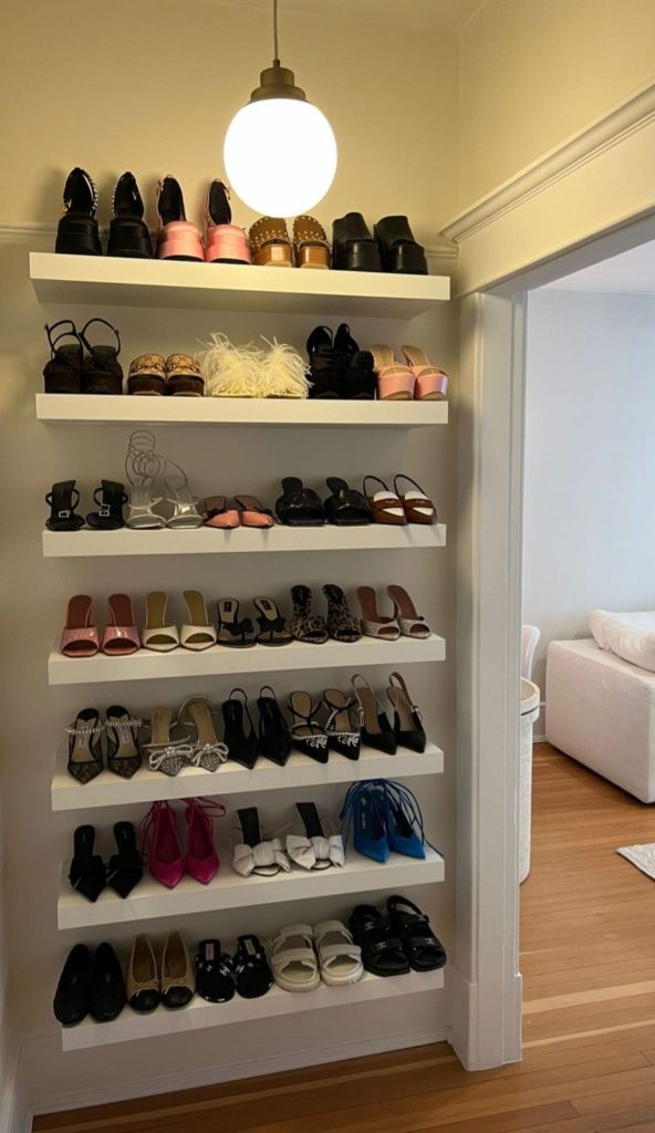 Seven white floating ledge shelves filling an alcove wall with rows of heels, sandals, and flats, lit by a glass globe pendant light.