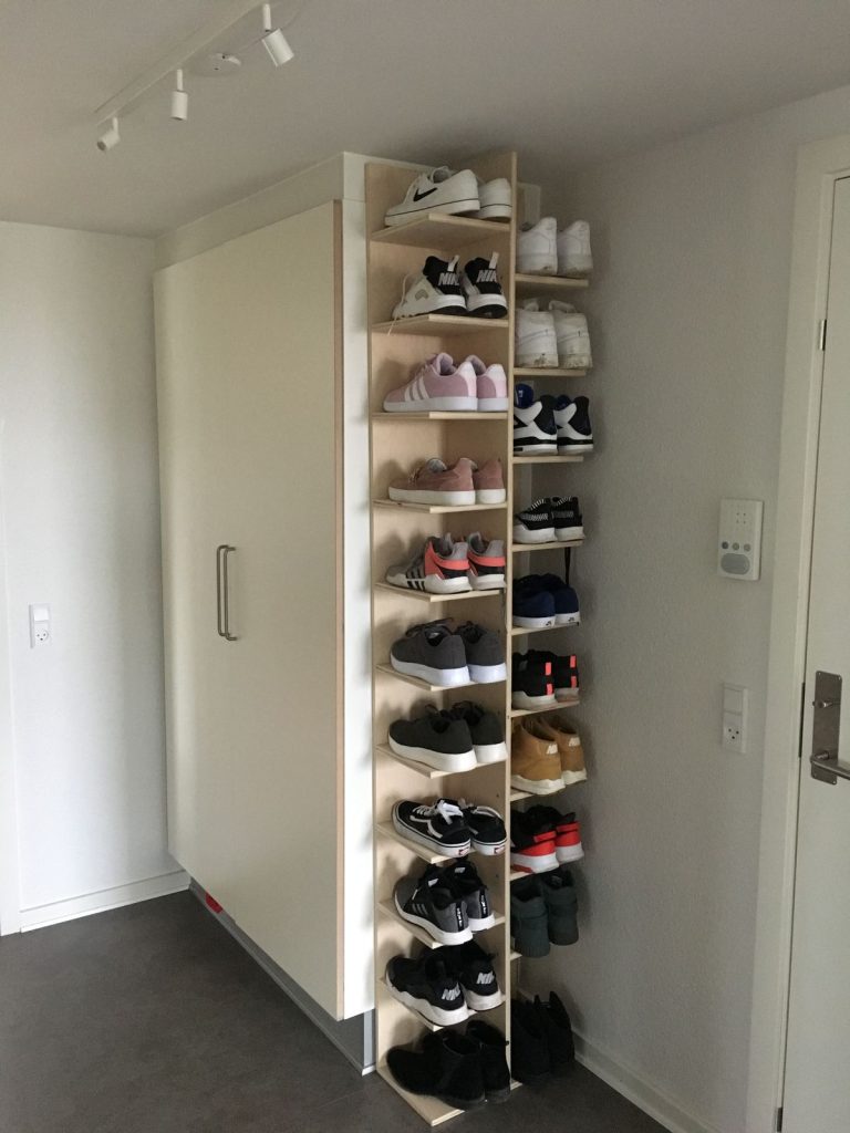 Tall narrow wooden shoe tower with staggered shelves against a white entryway wall, holding sneakers on both sides.