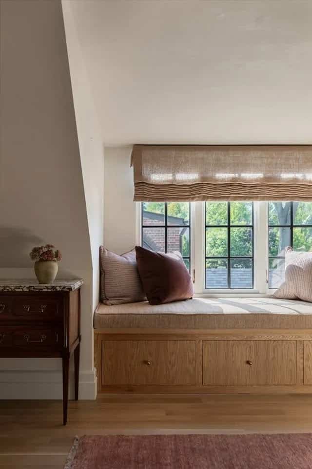 Window seat with a warm oak built in base and drawers, an oatmeal cushion, and dusty pink and burgundy pillows beneath a woven Roman shade and black framed windows.