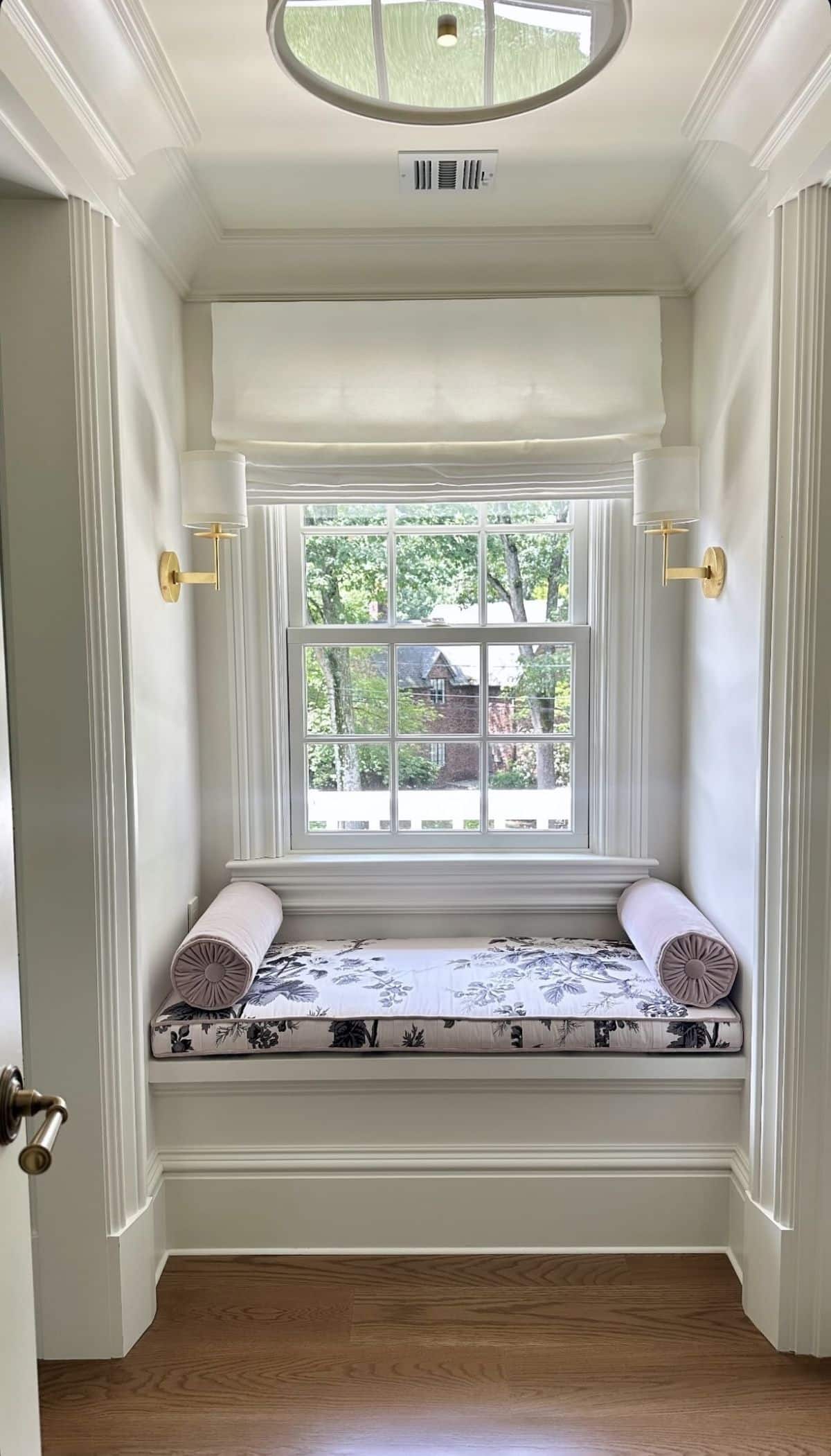 Formal window seat with a black and white floral chintz cushion and lilac velvet bolster pillows, framed by symmetrical brass sconces below an oval window.