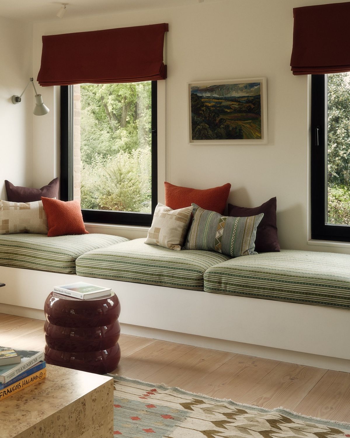 Long white built in window seat running beneath three black framed windows, topped with a green striped cushion and a mix of rust, plum, and patterned pillows.