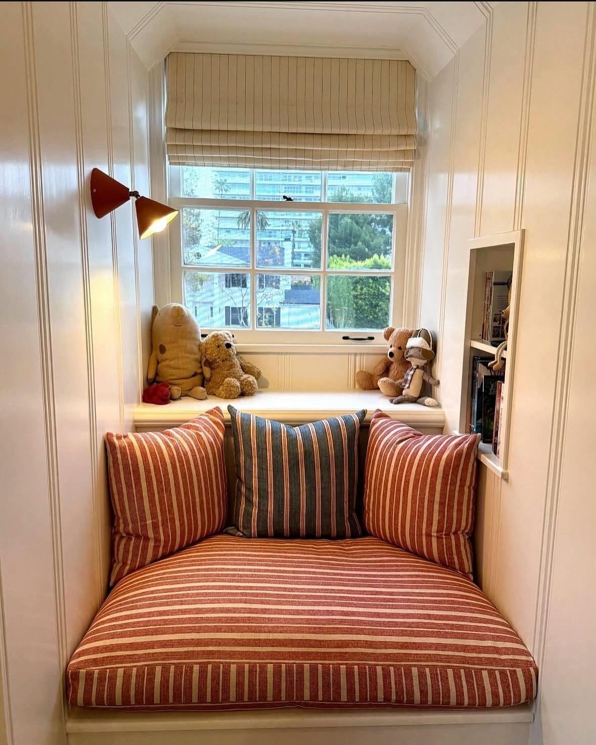 Cozy window seat nook set into a cream paneled alcove, with red and white striped cushions, a blue striped pillow, and a small built in bookshelf to the side.