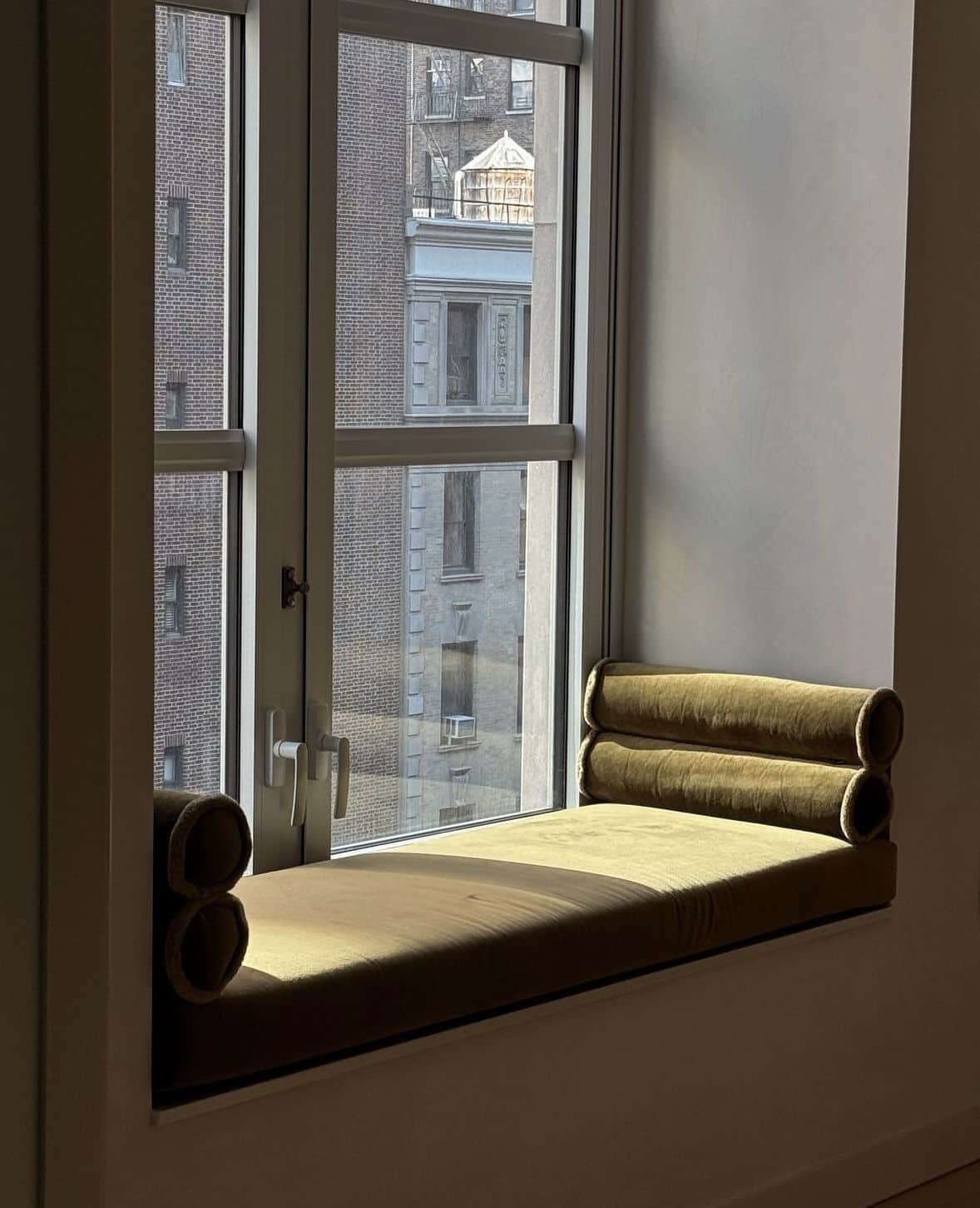 Modern window seat daybed upholstered in olive green velvet with rolled bolster arms, set beneath a black framed window overlooking a city view.