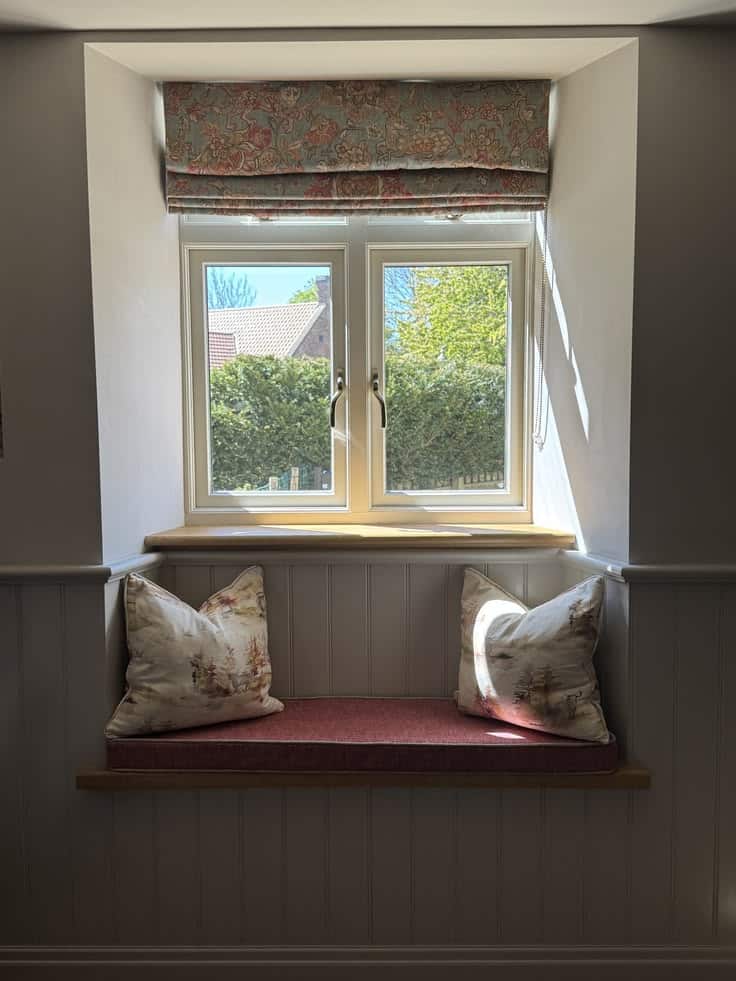 Cottage window seat recessed into a wall with grey tongue and groove paneling, a floral Roman shade, toile pillows, and a dusky pink seat cushion.