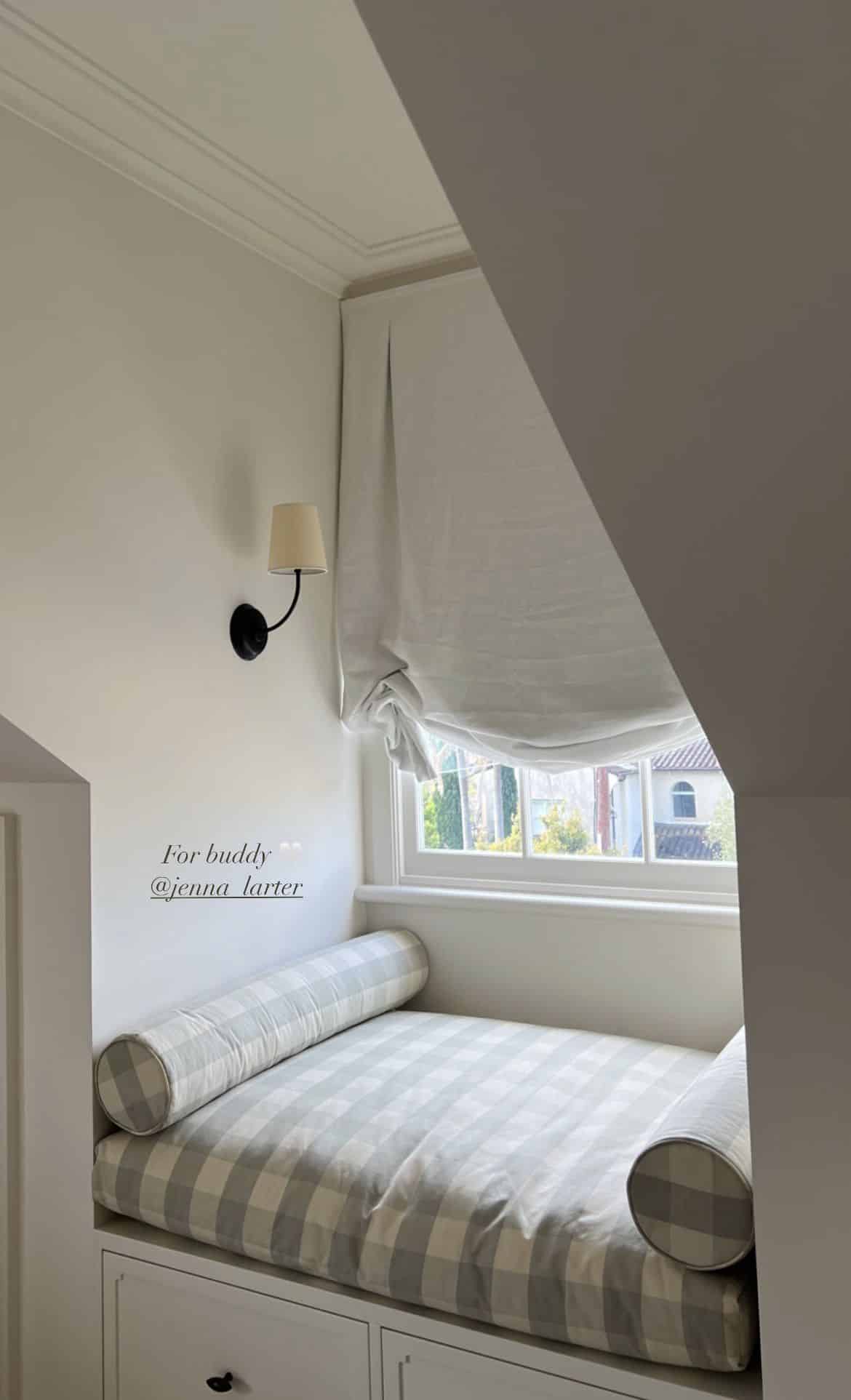 Window seat built into a sloped attic corner, upholstered in soft blue and white gingham with matching bolster cushions, beside a black wall sconce.
