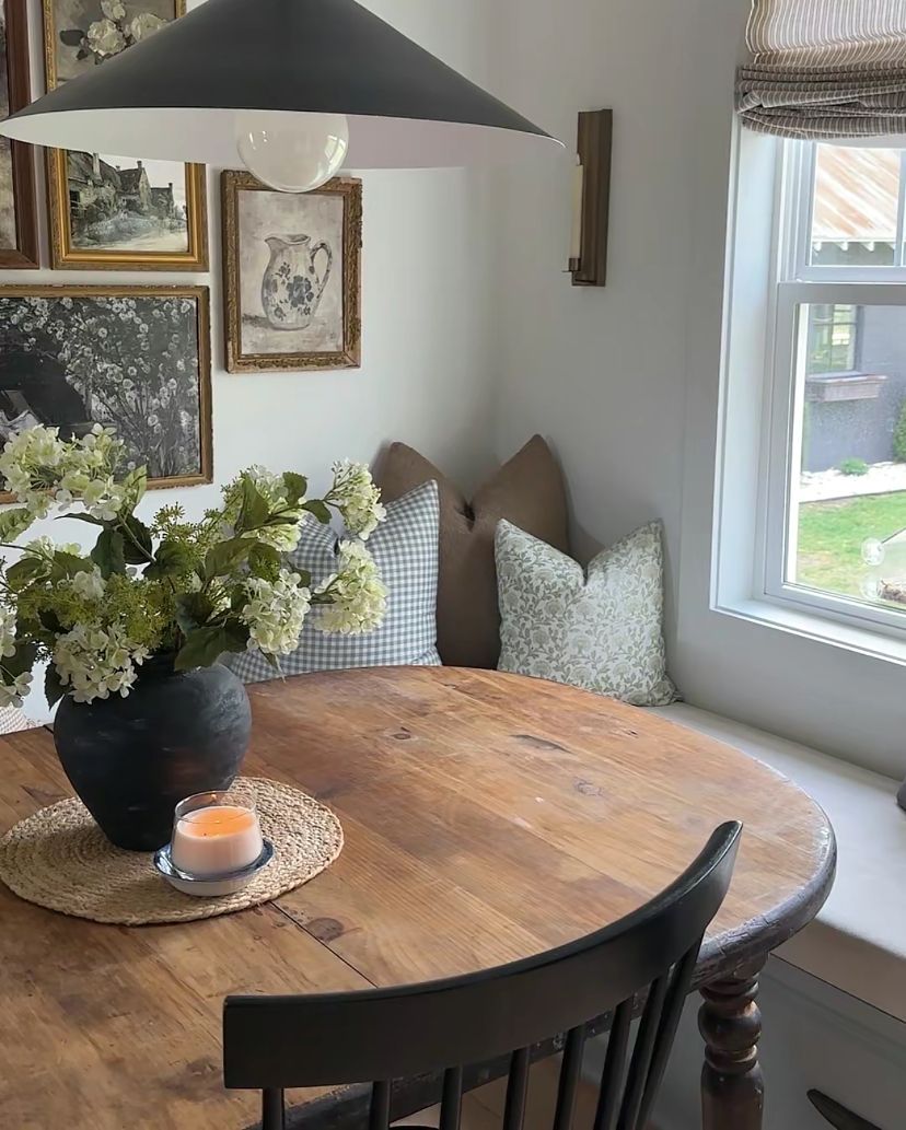 Breakfast banquette window seat with gingham and floral green pillows against a striped Roman shade, beside a round wood table and a vintage gallery wall.