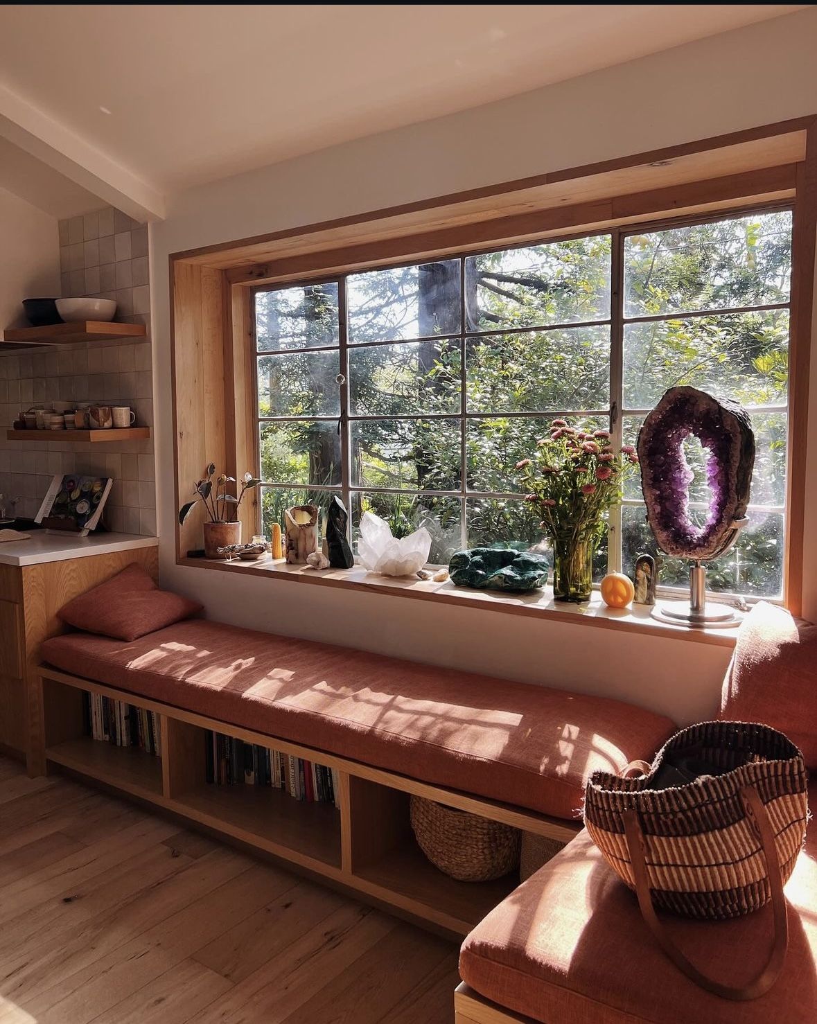 Boho window seat with a terracotta linen cushion and open wood shelving filled with books below, set beneath a large grid window lined with crystals and flowers.