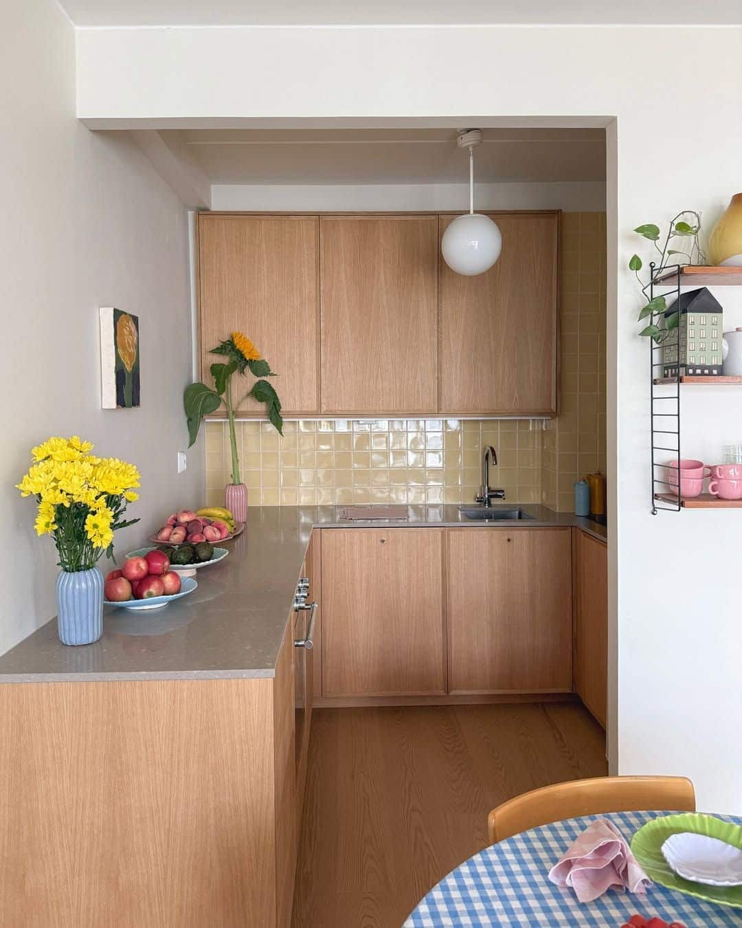 Light oak veneer kitchen with a pale butter yellow tile splashback, a wire string shelf holding pink mugs and plants, and an opal glass globe pendant.