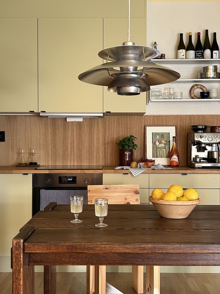 Mid century kitchen with soft butter cabinetry, a walnut splashback and benchtop, open shelving of glassware and wine, and a tiered metal pendant over a timber dining table.