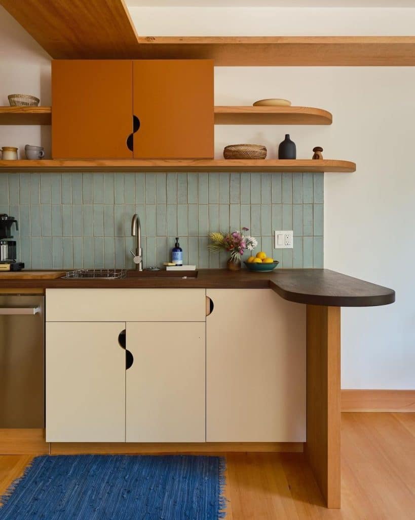 Two-tone kitchen with warm orange upper cabinets, cream lower cabinets, half-moon cut-out pulls, a sage green stacked tile splashback and a curved walnut breakfast bar.