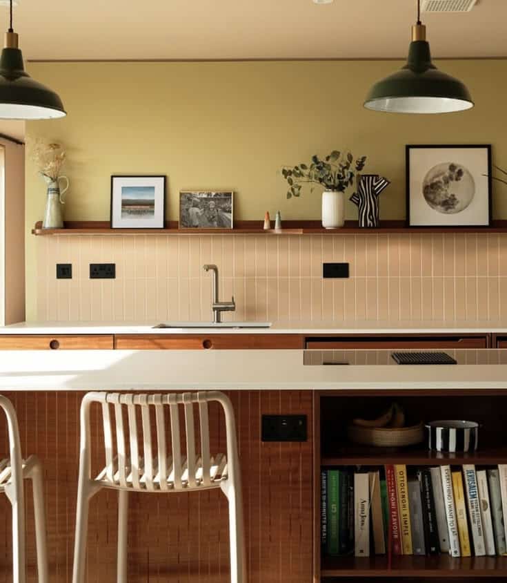 Colourful kitchen with a chartreuse feature wall, a blush stacked tile splashback, deep green dome pendants, walnut cabinetry and a breakfast bar lined with cookbooks.