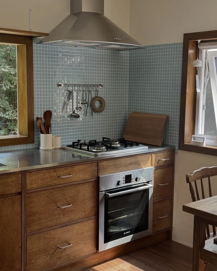 Warm stained timber kitchen with slim silver bar pulls, a dusty blue mosaic tile splashback, a stainless benchtop and chimney hood, and a utensil rail.