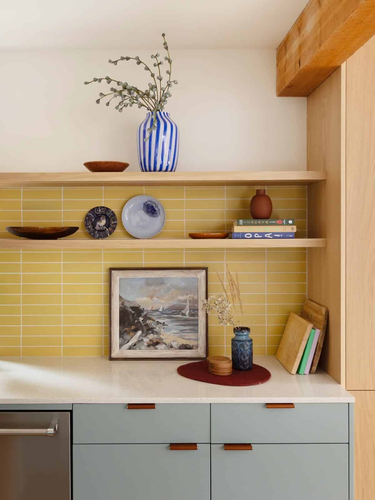 Kitchen detail with a mustard stacked tile splashback, sage green cabinets with tan leather tab pulls, a speckled benchtop, oak shelves and a striped vase.