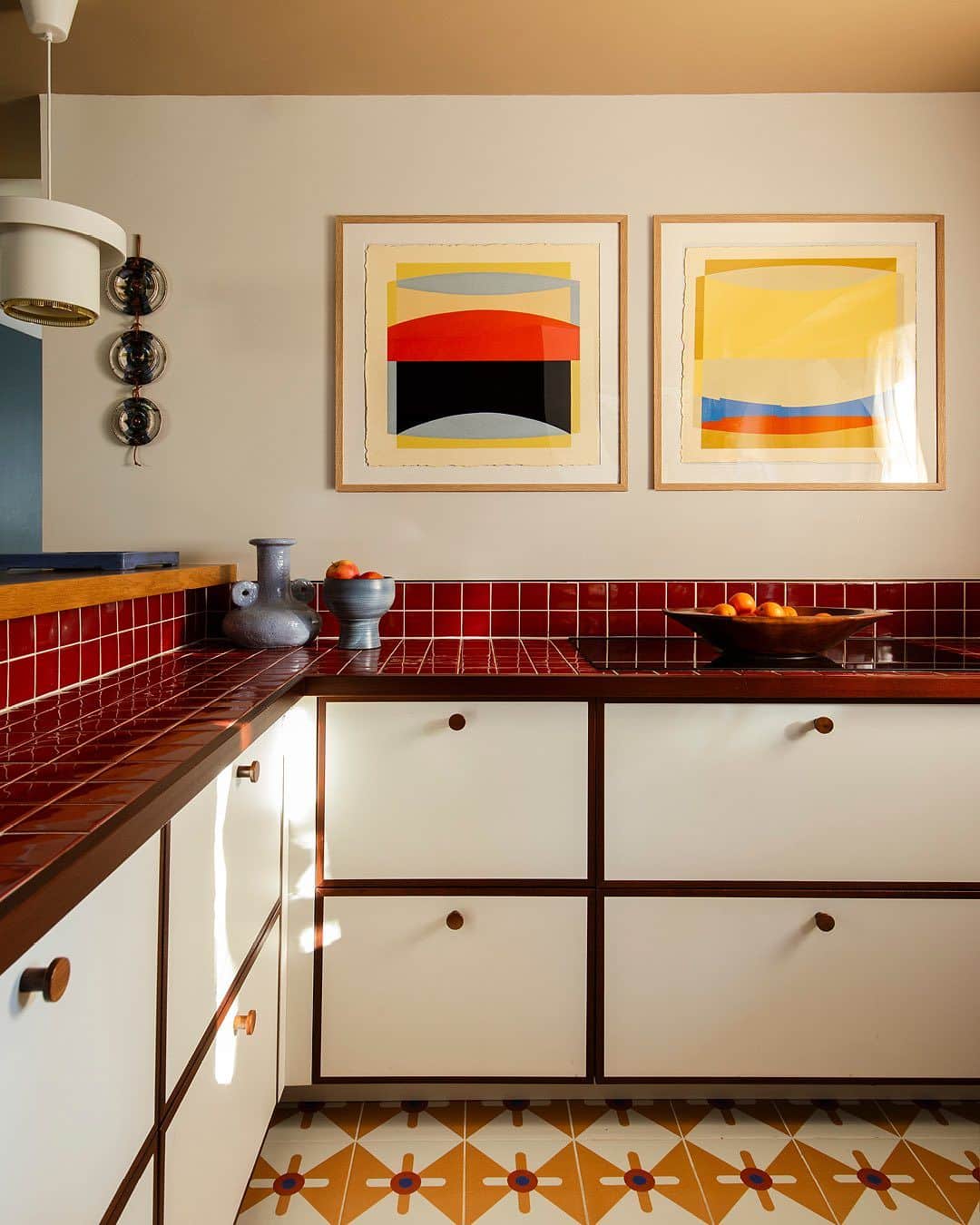 Bold retro kitchen with deep red glossy tiled benchtops, white cabinets with dark reveals and timber knobs, a star-patterned encaustic floor and geometric art prints.