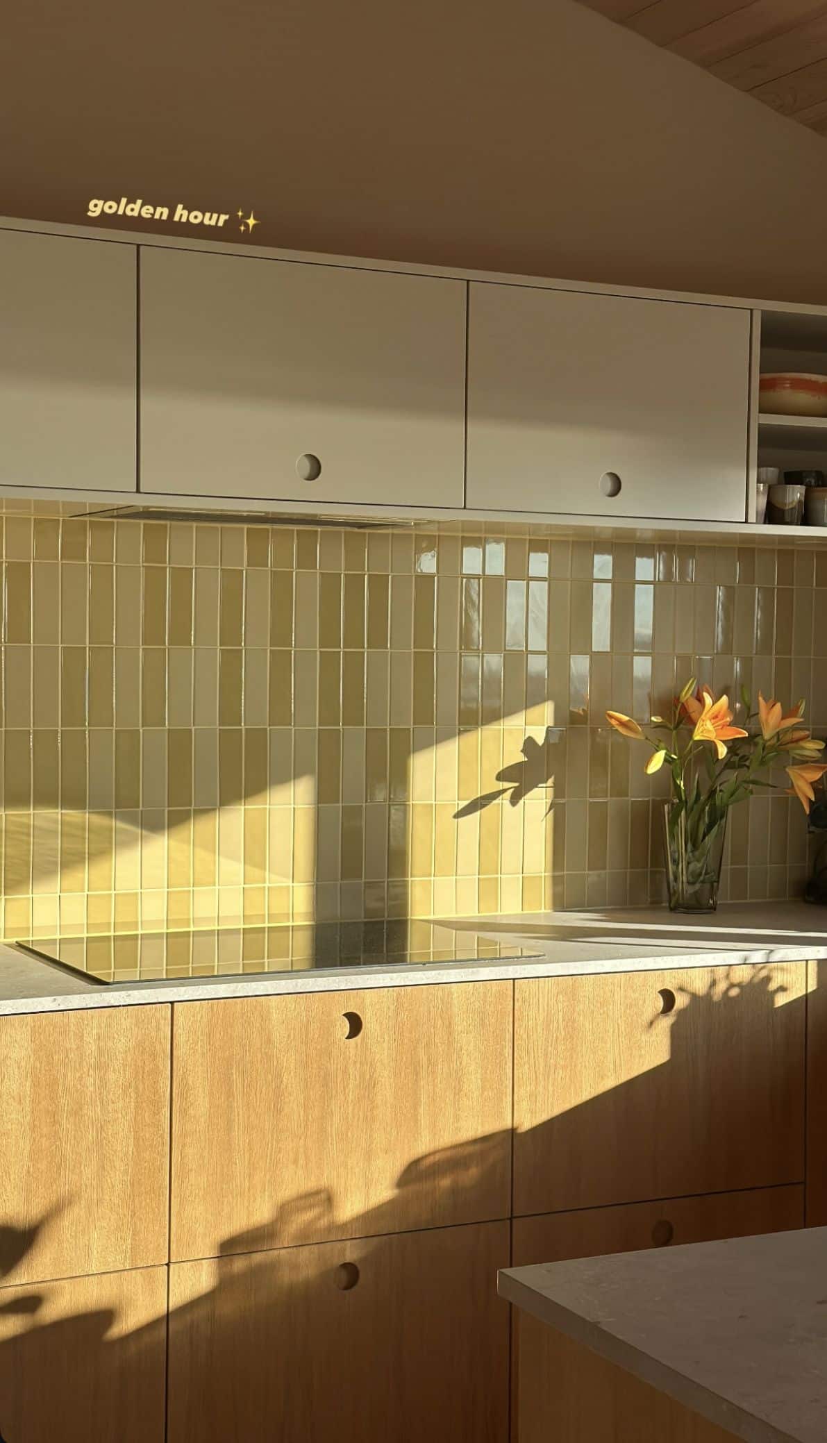 Warm oak kitchen at golden hour with circular cut-out cabinet pulls, an olive stacked tile splashback, greige upper cabinets and a stone benchtop.