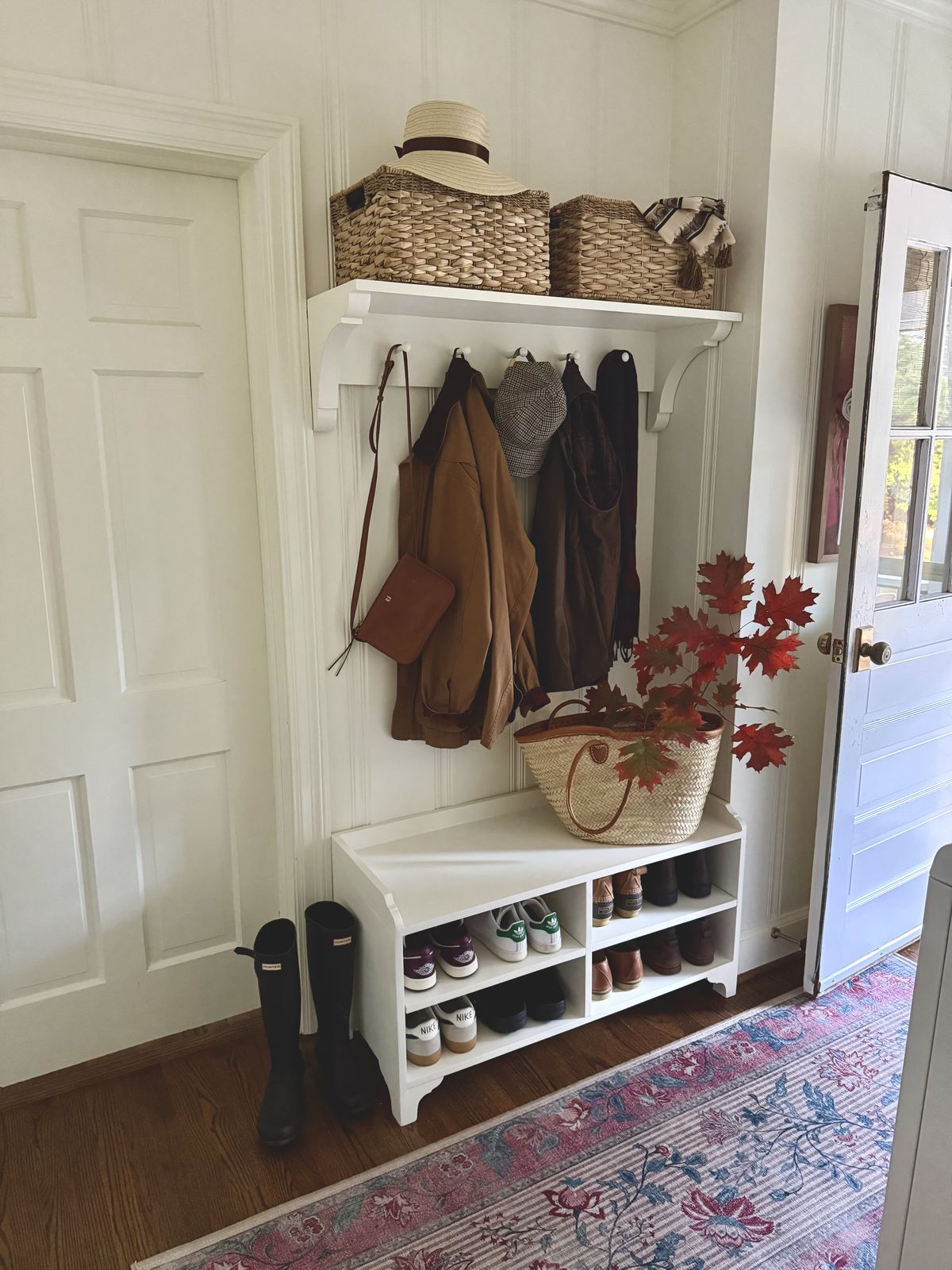 White board and batten entryway with a shelf and hooks holding coats and hats, a straw hat and woven baskets on top, and a white storage bench with open cubbies full of shoes beside black rain boots.