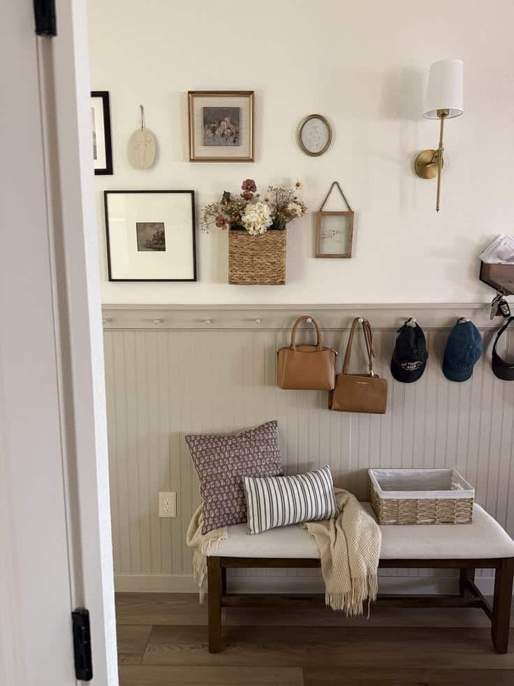 Neutral greige beadboard entry with a peg rail holding handbags and caps, a mail organizer, a layered gallery wall of framed art and a basket of dried florals, and an upholstered bench with pillows.