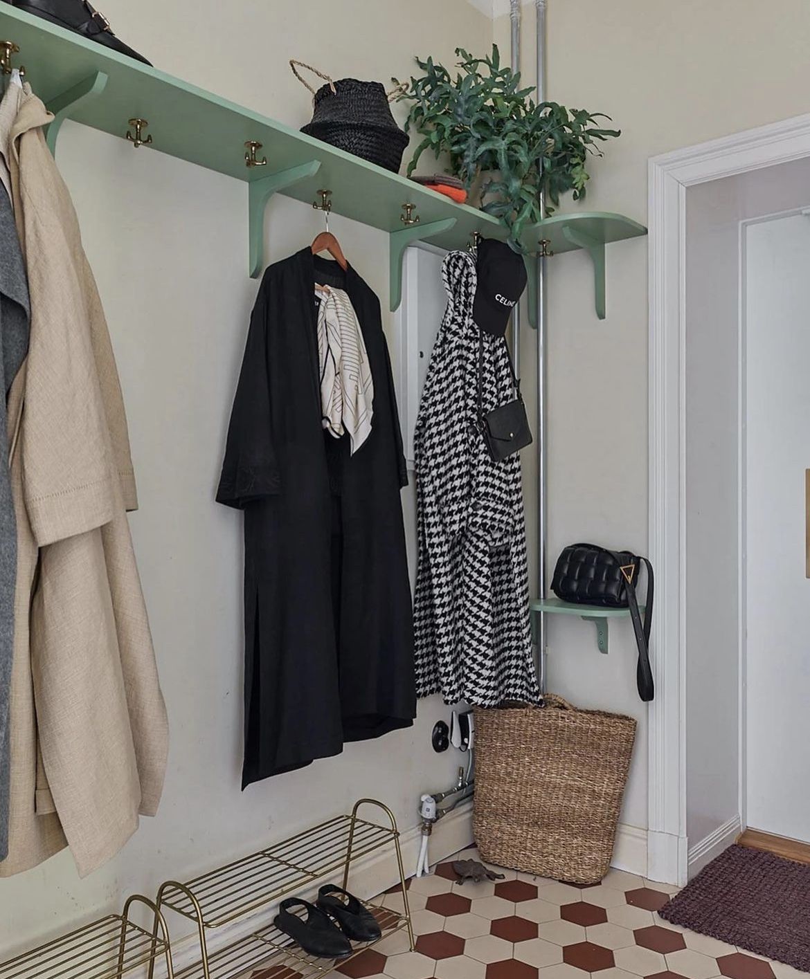 Eclectic entry with mint green painted shelf-and-hook rails holding coats and a bag, a trailing plant and basket on the upper shelf, a brass wire shoe rack, and a red and white checkerboard tile floor.