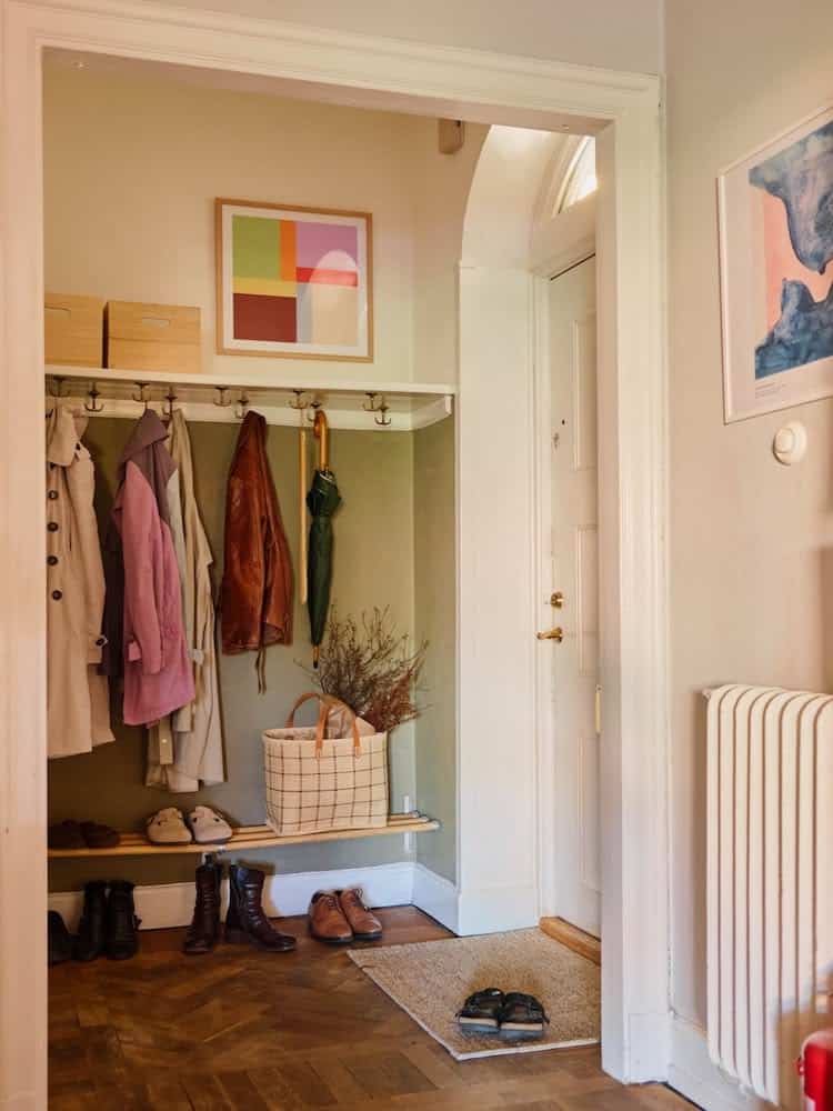 Shallow recessed alcove painted olive green, with a ledge of brass hooks holding trench coats and jackets, framed abstract art above, and a slim wood bench with shoes underneath and a checked tote on top.