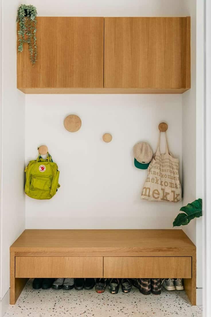 Modern white and oak mudroom nook with wall-mounted oak upper cabinets, round wooden peg hooks holding a backpack and tote, and an oak floating bench with drawers over a row of shoes.