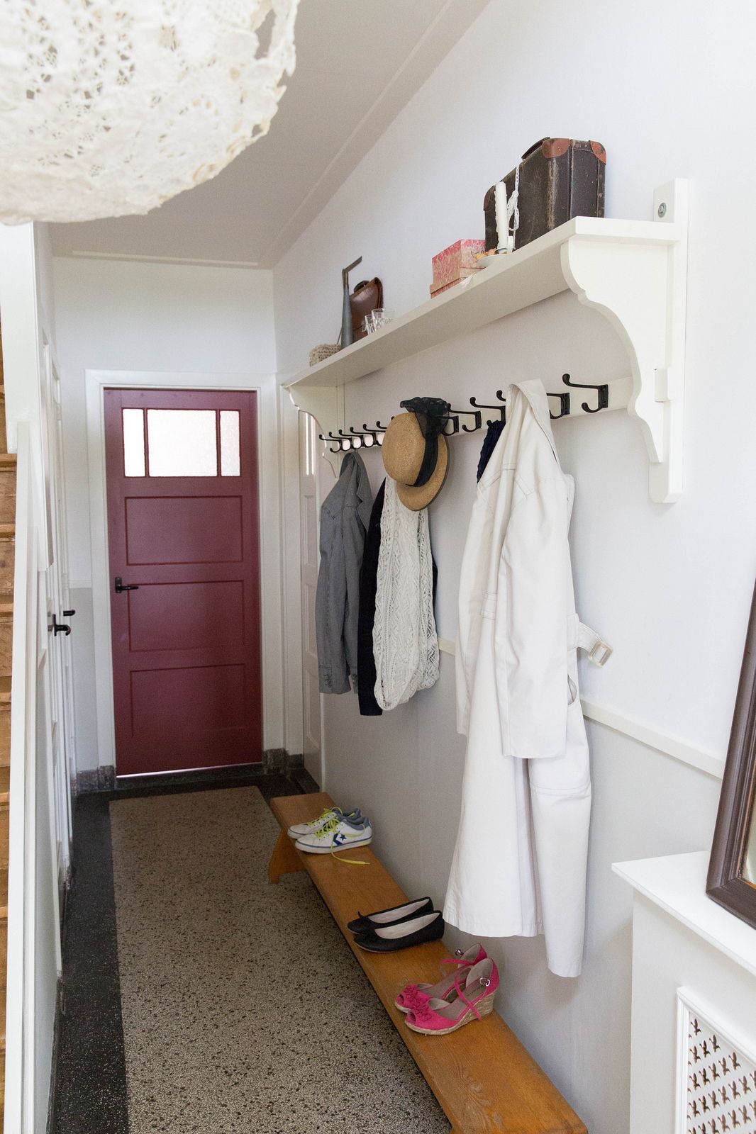Narrow hallway entry with a white shelf and a long black cast-iron hook rail holding a shirt, hat, scarf, and trench coat, vintage suitcases styled on the shelf, and a long low wood bench with shoes.