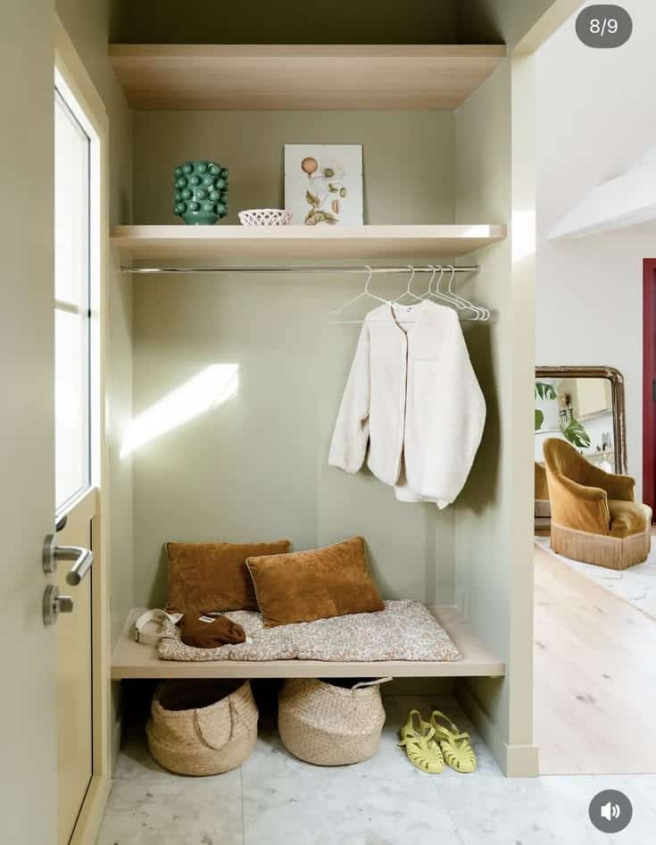 Sage green mudroom alcove with a light oak floating shelf, a hanging rail with a sherpa jacket on hangers, a styled shelf above, a cushioned platform bench with pillows, and seagrass basket bags underneath.