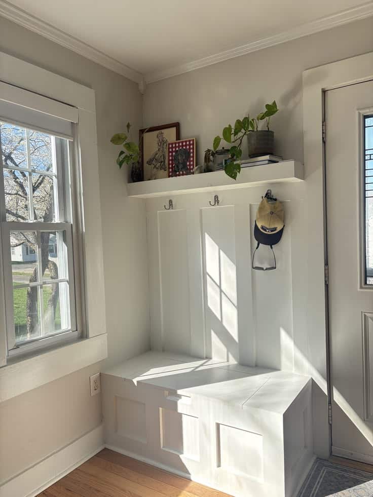 Bright corner entry with warm white board and batten, two metal hooks holding a cap, a floating shelf styled with trailing plants and a framed print, and a white paneled Craftsman-style built-in storage bench.