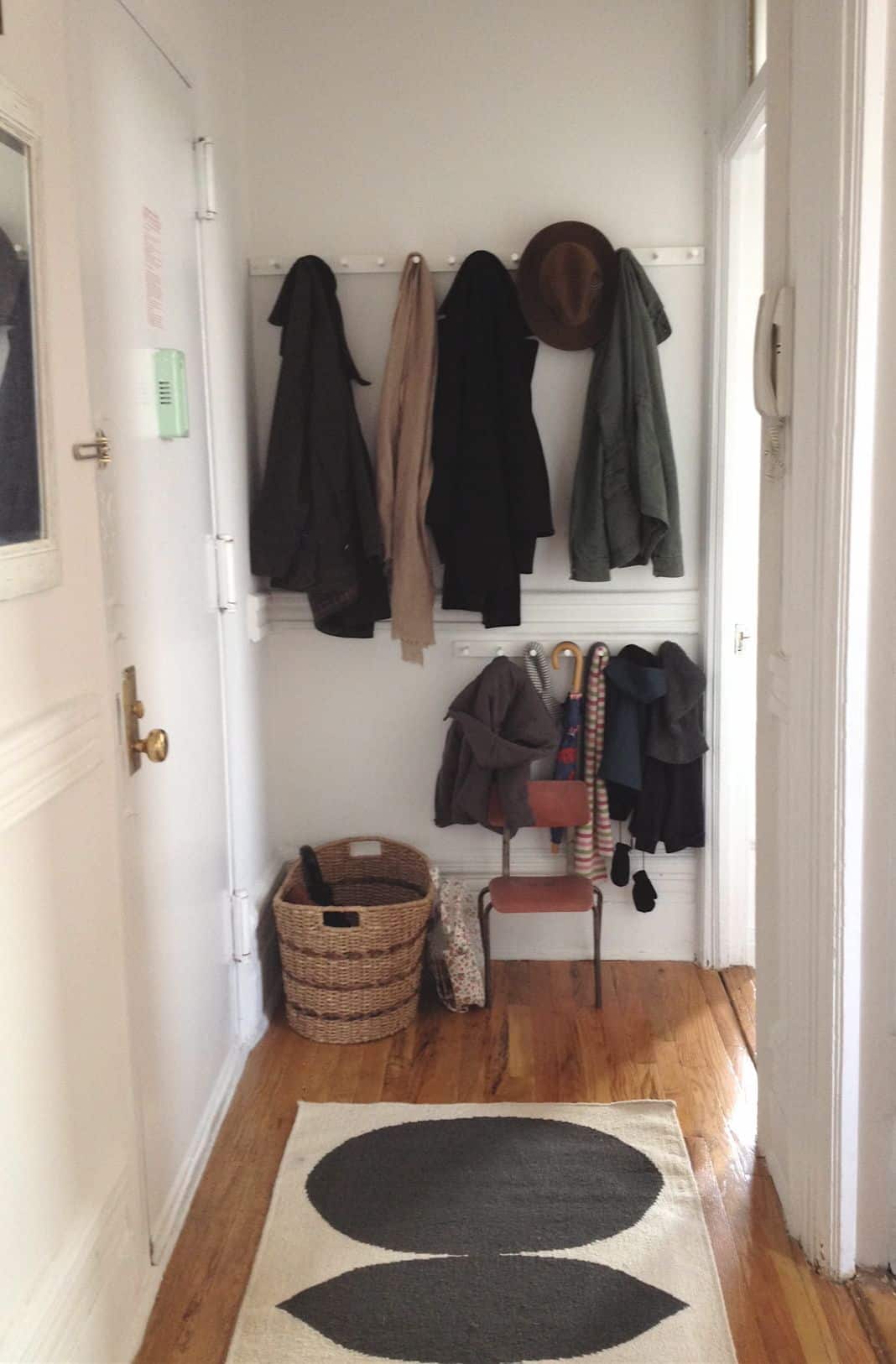 Minimalist apartment entry with two rows of white peg rails holding dark coats, a scarf, a felt hat, and a green jacket, a small vintage rust-orange chair, a basket of umbrellas, and a graphic circle rug.