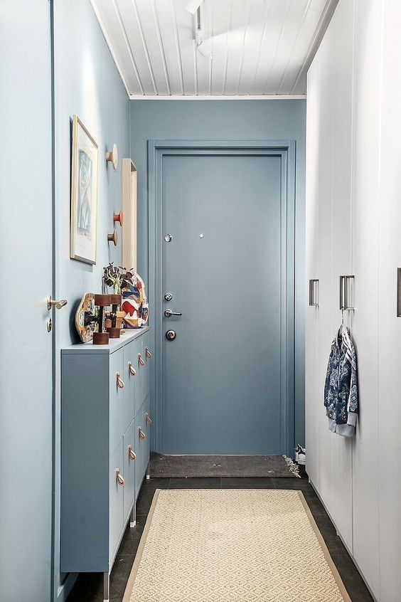 Narrow dusty blue hallway entry with a slim blue tilt-out shoe cabinet with rattan pulls, wooden pegs and a red hook on the wall, framed art, a white paneled wardrobe opposite, and a patterned runner on a slate floor.