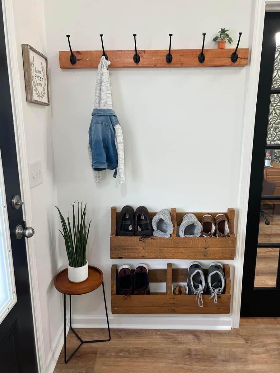 Rustic reclaimed wood hook rail with black hooks holding a denim jacket and hoodie, above two wall-mounted wooden slat shoe racks, next to a small round side table with a potted snake plant.