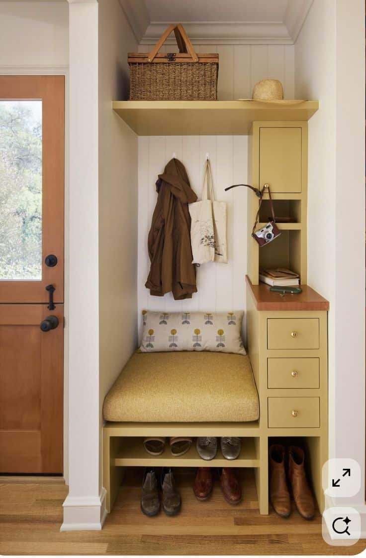 Custom mudroom nook painted mustard yellow with a cushioned bench, an open shoe shelf below, two hooks on a white shiplap wall, and a narrow cabinet with drawers and an open cubby beside it.