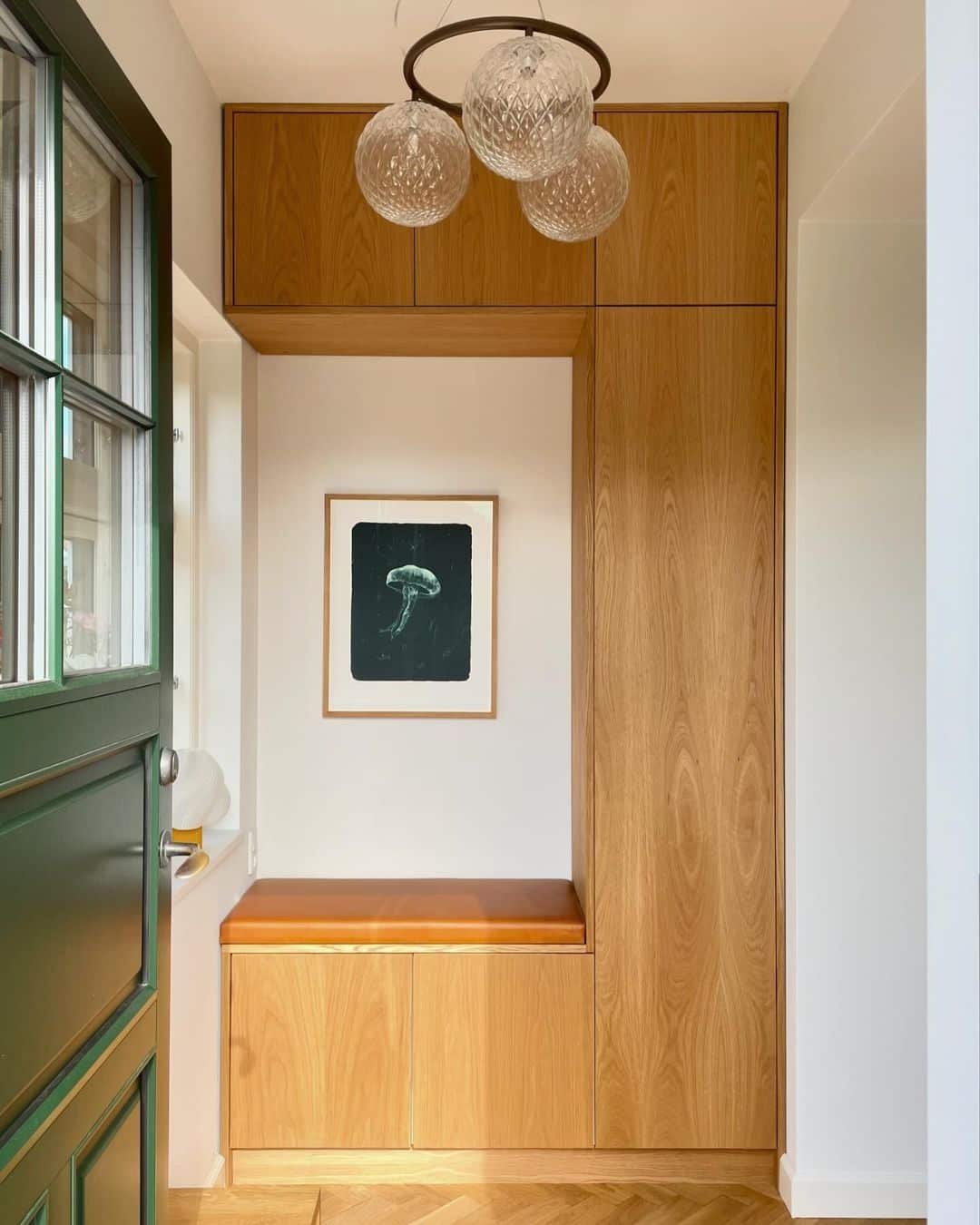 Mid-century entryway with warm oak floor-to-ceiling cabinetry, an integrated bench with a tan leather cushion, a framed print, a cluster of glass globe pendant lights, and a green glass-paned door.
