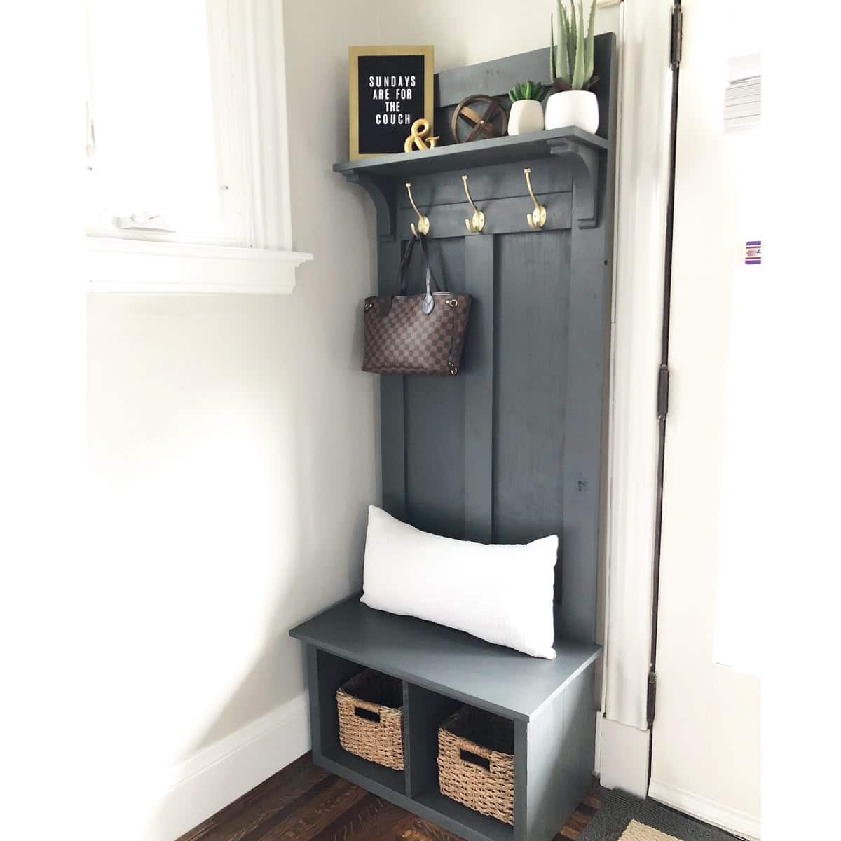 Elegant cream paneled entryway with three antique brass double hooks holding a cap, a canvas tote, and a blazer, a framed vintage print above, and a rustic wooden stool with riding boots beside it.