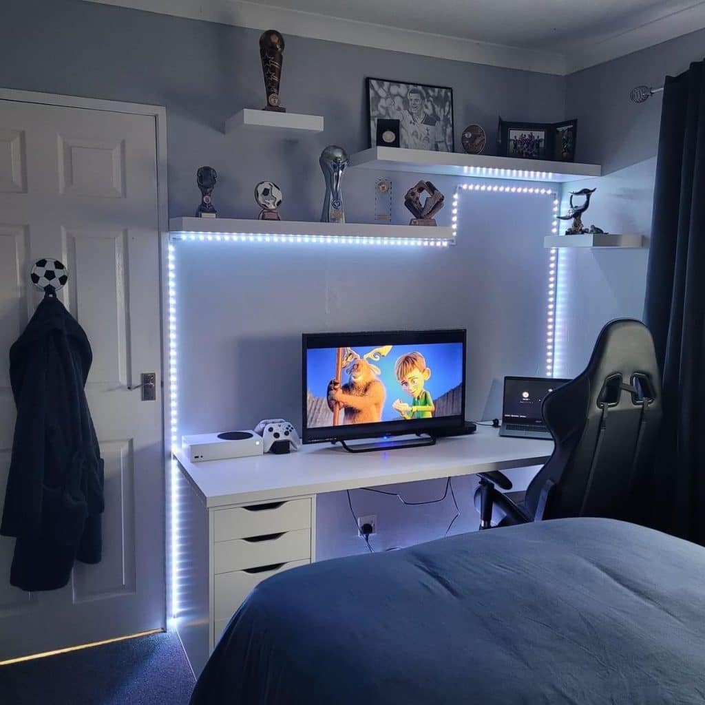 Teen bedroom with pale gray walls, white floating shelves holding trophies and framed photos, white LED strip lights outlining the shelves and desk wall, a white desk with drawers, a TV and gaming console, and a black gaming chair.