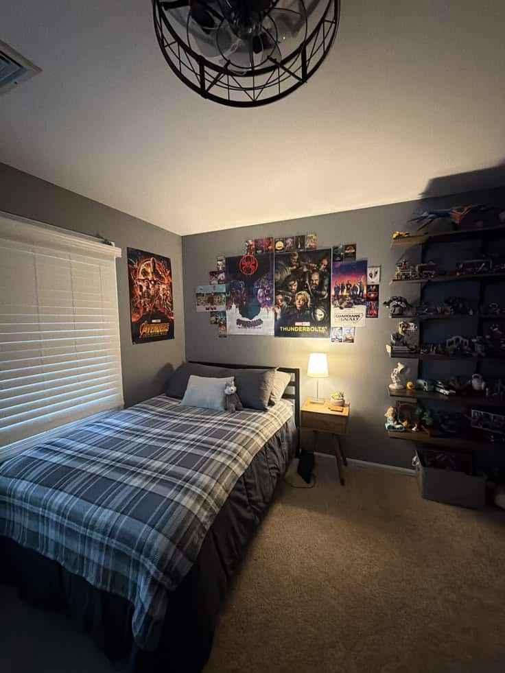Teen boys bedroom with gray walls, a collage of superhero movie posters above the bed, blue and gray plaid bedding, a small wooden nightstand with a lamp, and dark floating shelves filled with figures and models.