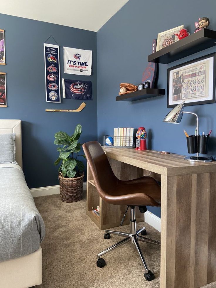 Teen bedroom with slate blue walls, hockey team banners and a mini hockey stick on the wall, a rustic wood desk with a brown leather swivel chair, black floating shelves with sports memorabilia, and a potted plant in a woven basket.