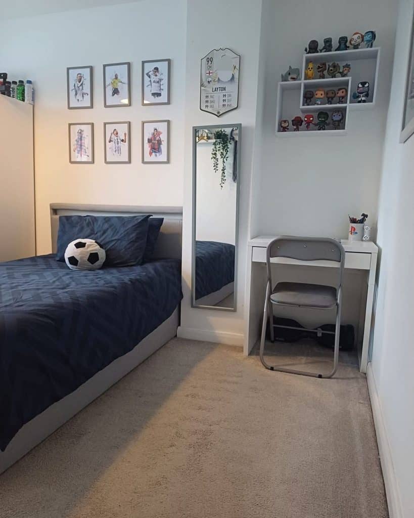 Teen boys bedroom with white walls, a grid of six framed soccer player prints above a gray upholstered bed with navy bedding, a tall floor mirror, a small white desk with a folding chair, and a white shelf unit of collectible figures.